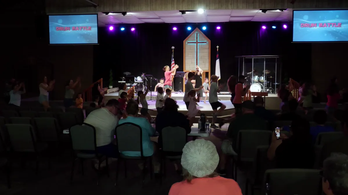 An evening program — children on the stage in front of a lit cross, families watching from tables and chairs.