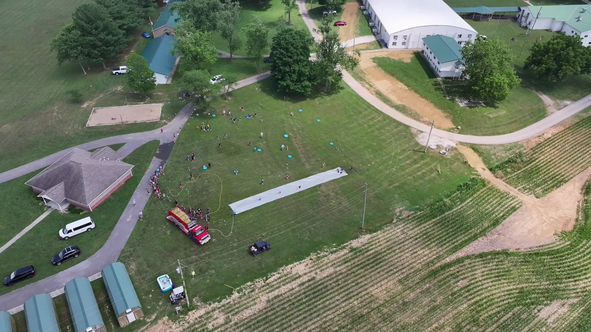 The camp field from above on a water-game day — a long sheet of plastic down the slope, a fire truck feeding the hose, dorms and cornfields beyond.