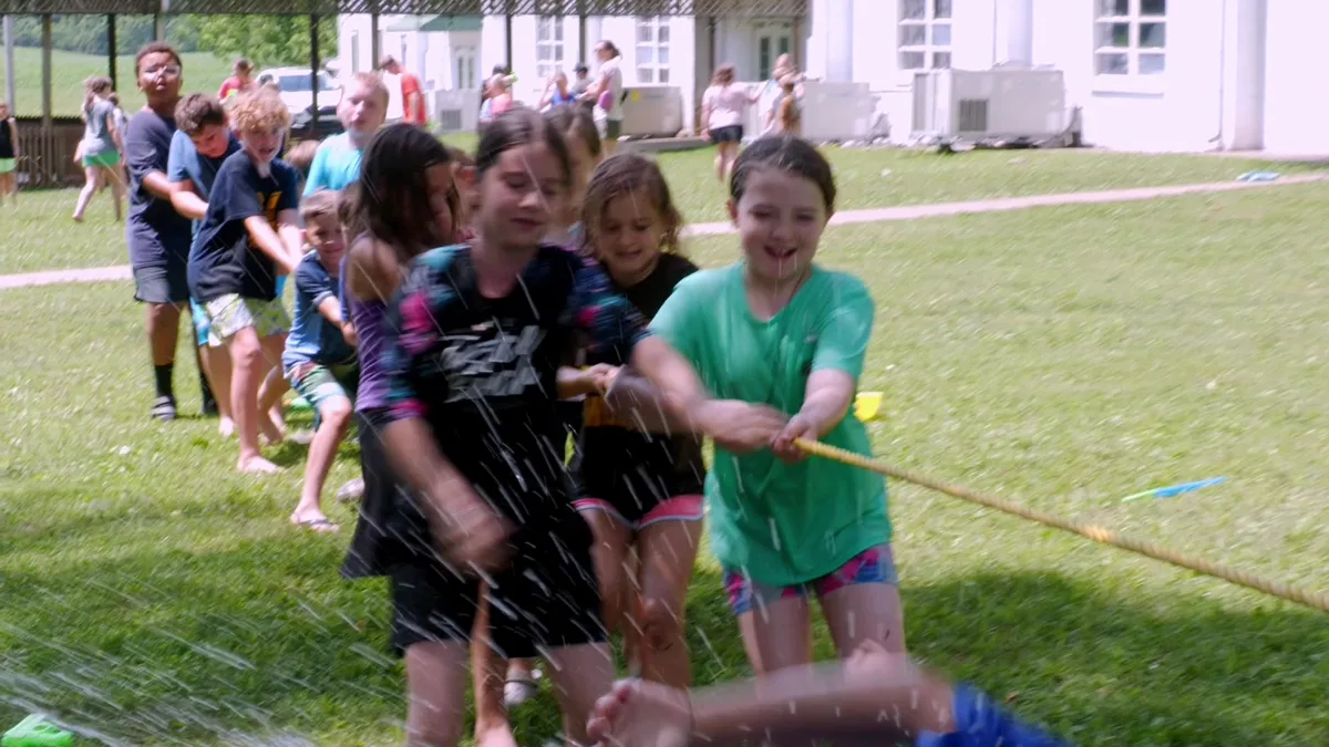 Campers hauling on a tug-of-war rope through a spray of water, soaked and laughing.