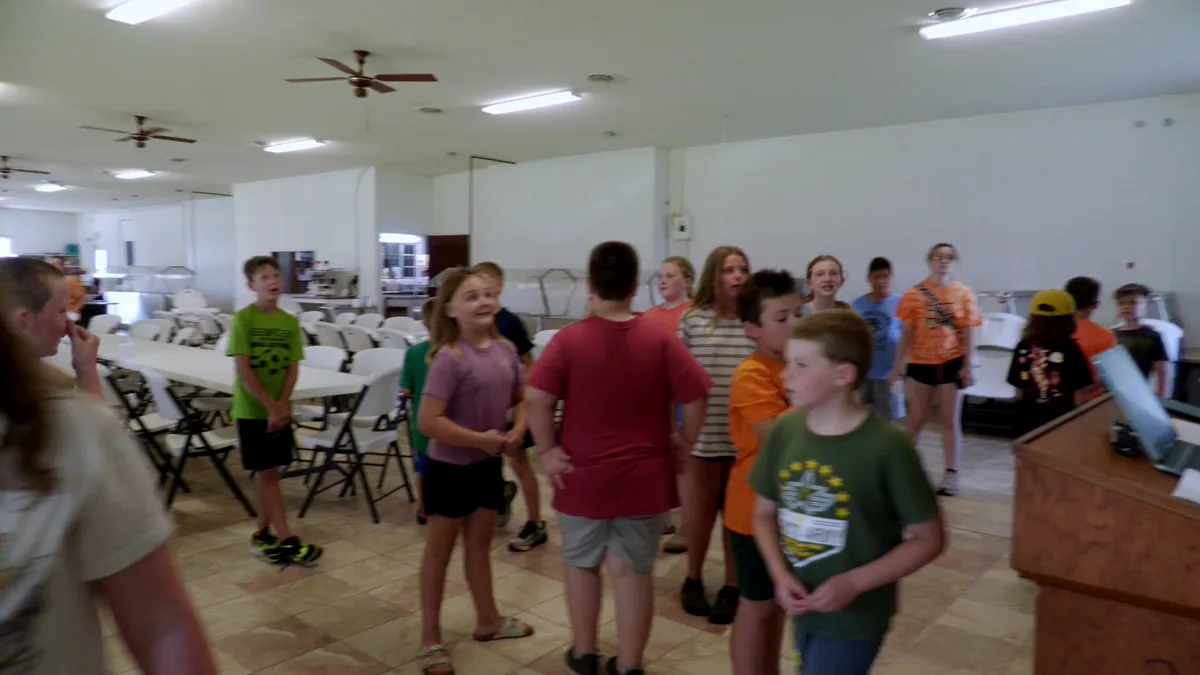 Inside the dining hall — long folding tables, ceiling fans and the serving counter, campers crossing the tiled floor.