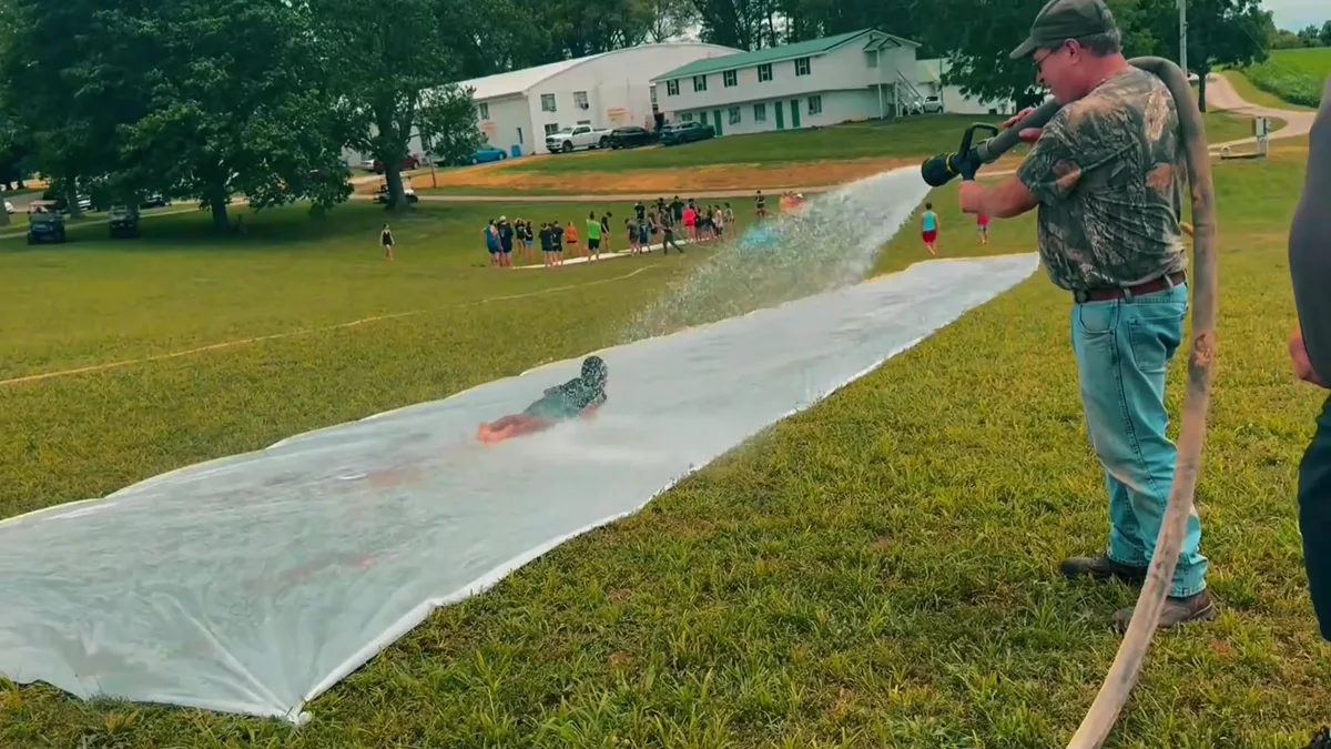 A member of staff playing a fire hose over the plastic sheeting as a camper slides down it, the dorms behind.