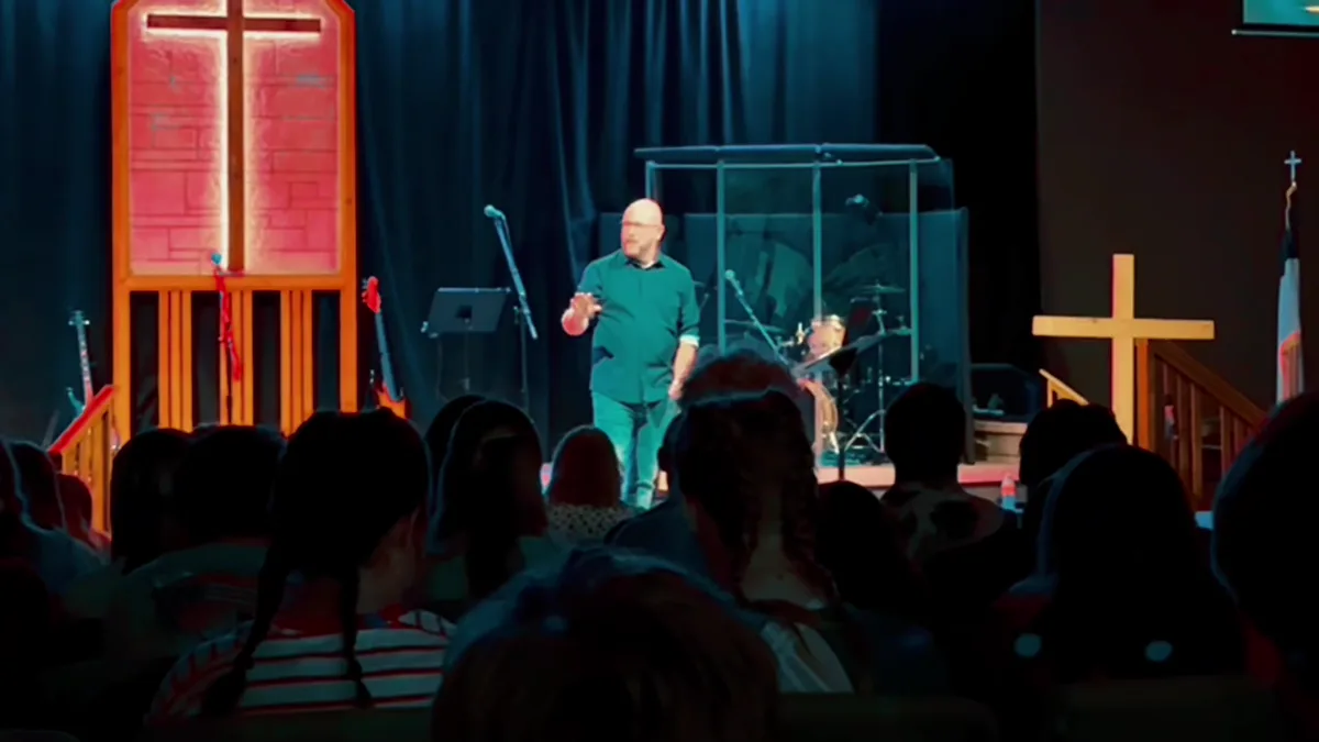 A speaker mid-message in front of a lit cross, teenagers watching from their seats.
